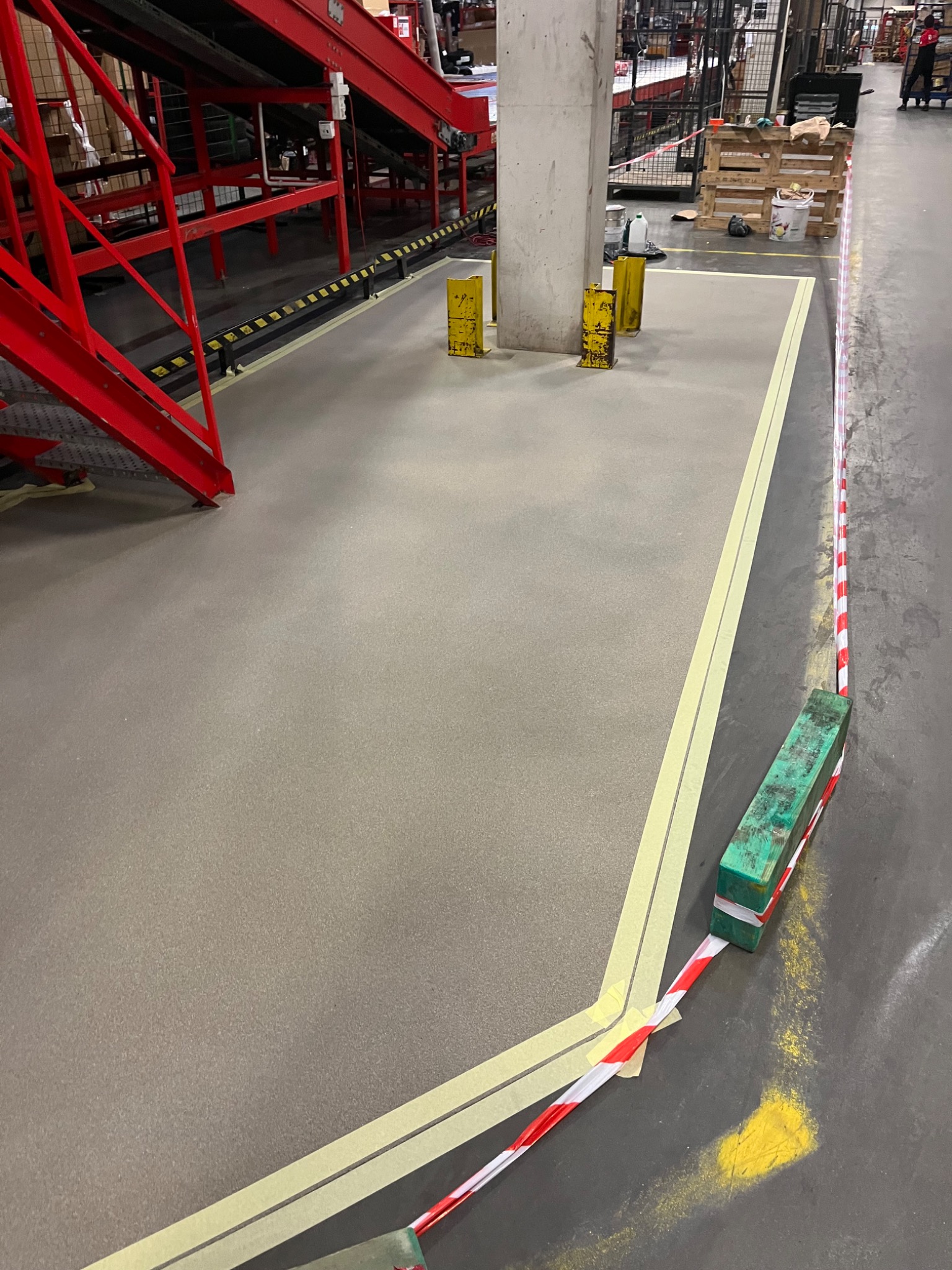Szara, gładka posadzka w hali przemysłowej, wyklejona taśmą malarską i zabezpieczona taśmą ostrzegawczą, widoczne słupy ochronne i fragment konstrukcji schodów.