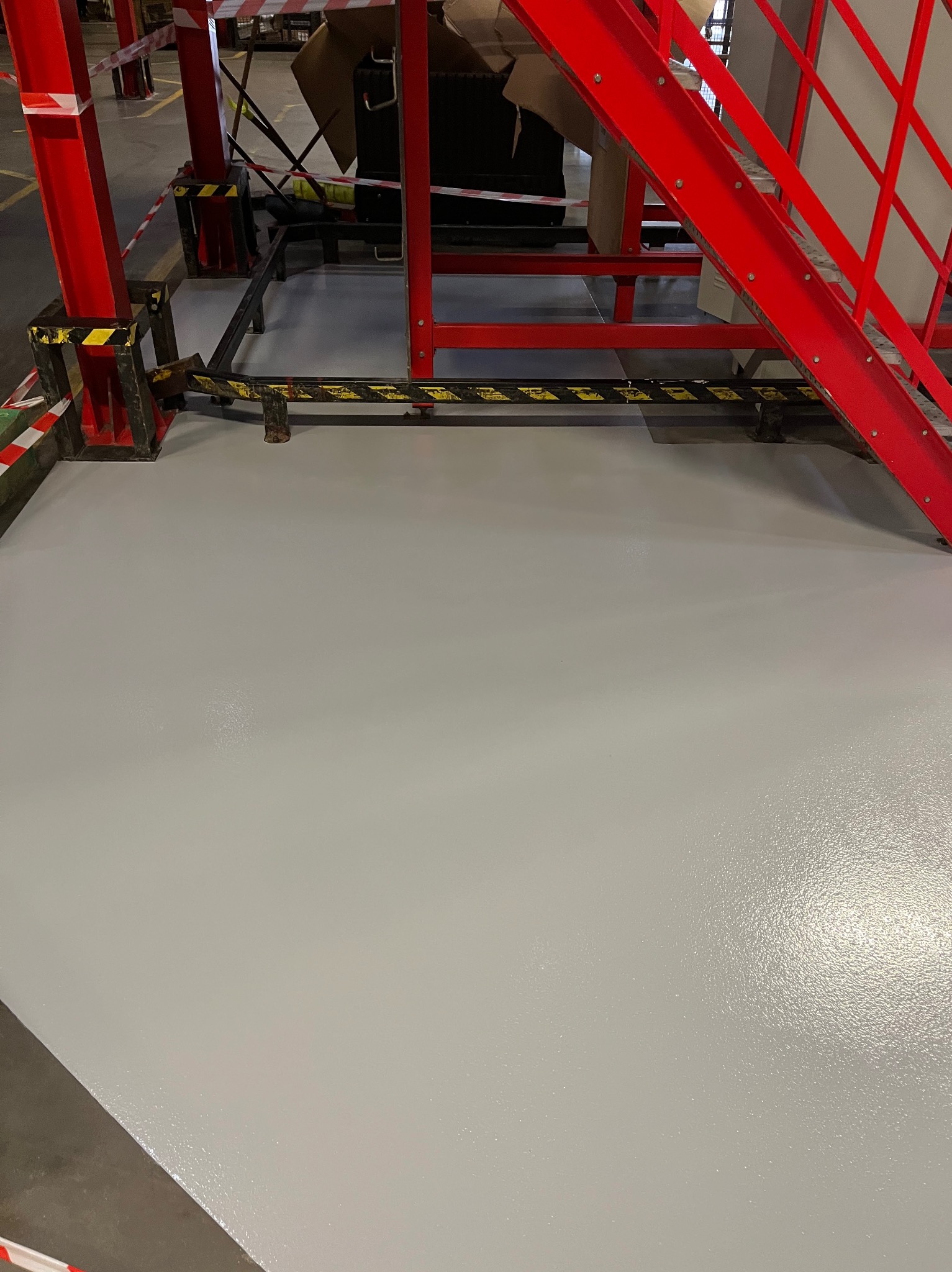 Jasnoszara, gładka posadzka w hali przemysłowej, z widocznymi elementami konstrukcji stalowej w kolorze czerwonym i żółto-czarnymi pasami ostrzegawczymi.