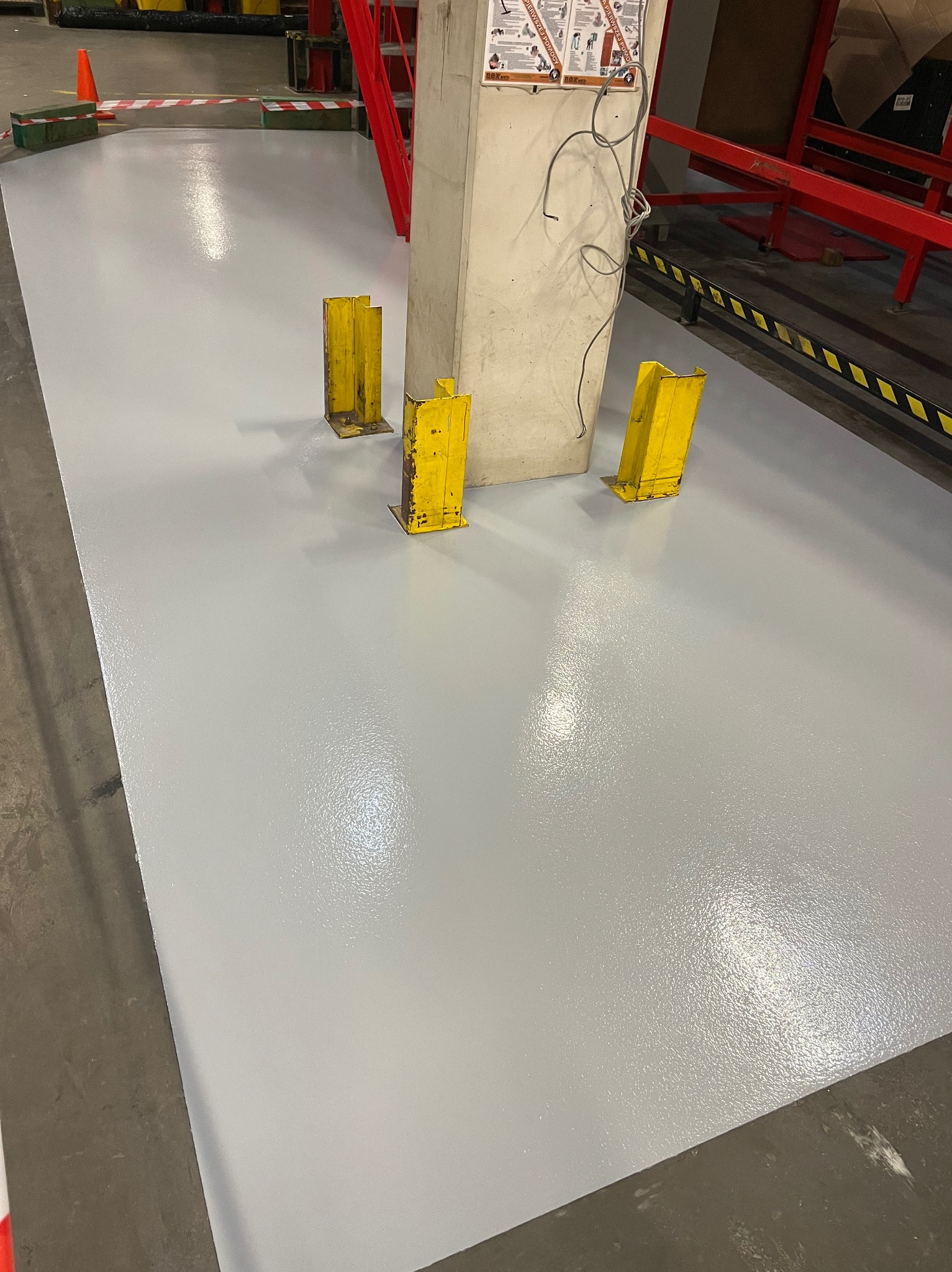 Jasnoszara posadzka żywiczna w hali produkcyjnej, z żółtymi osłonami słupa i widocznymi elementami konstrukcyjnymi hali.