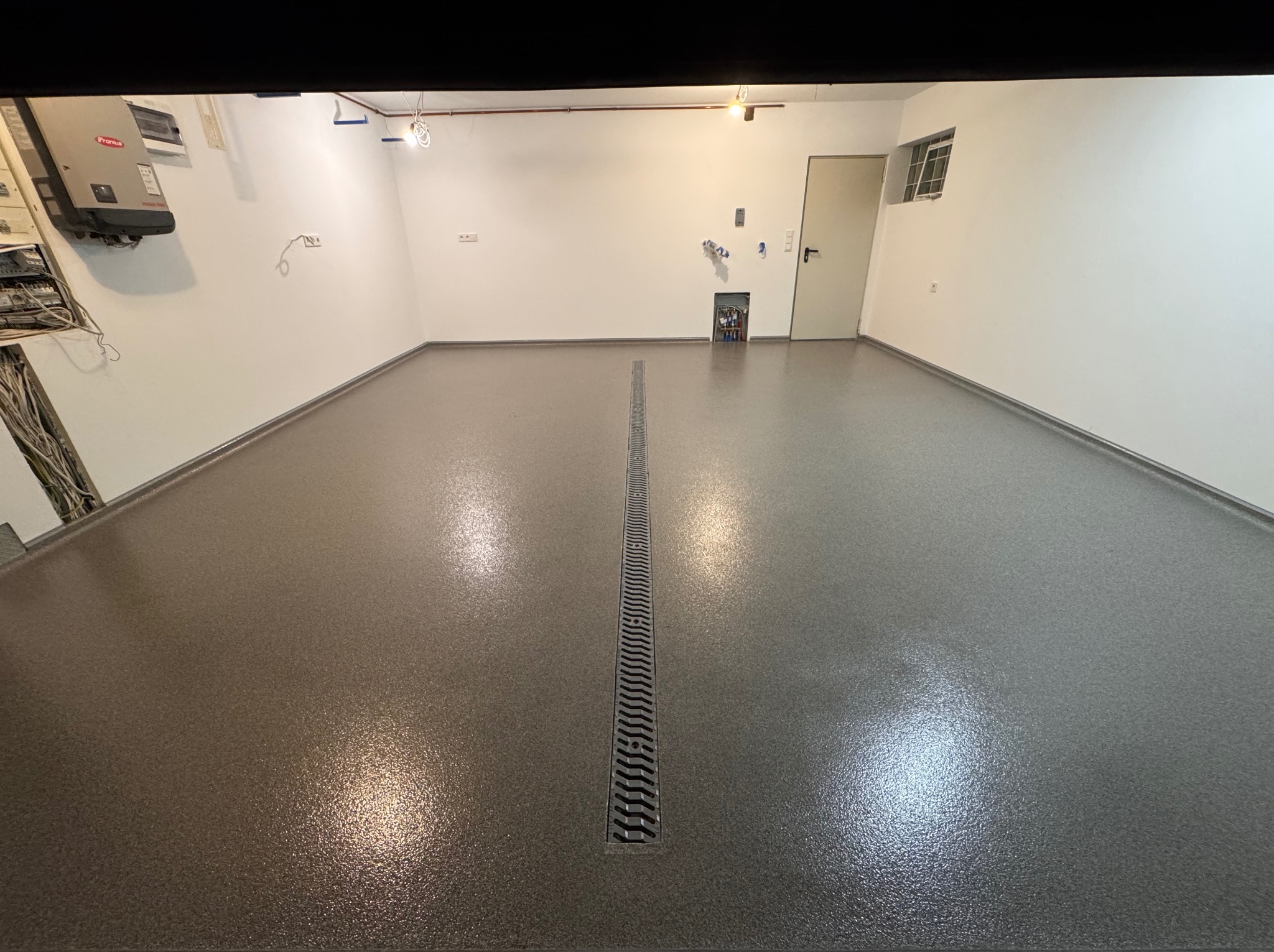 Szara, błyszcząca posadzka z odpływem liniowym w garażu lub pomieszczeniu gospodarczym, widoczne instalacje na ścianach.