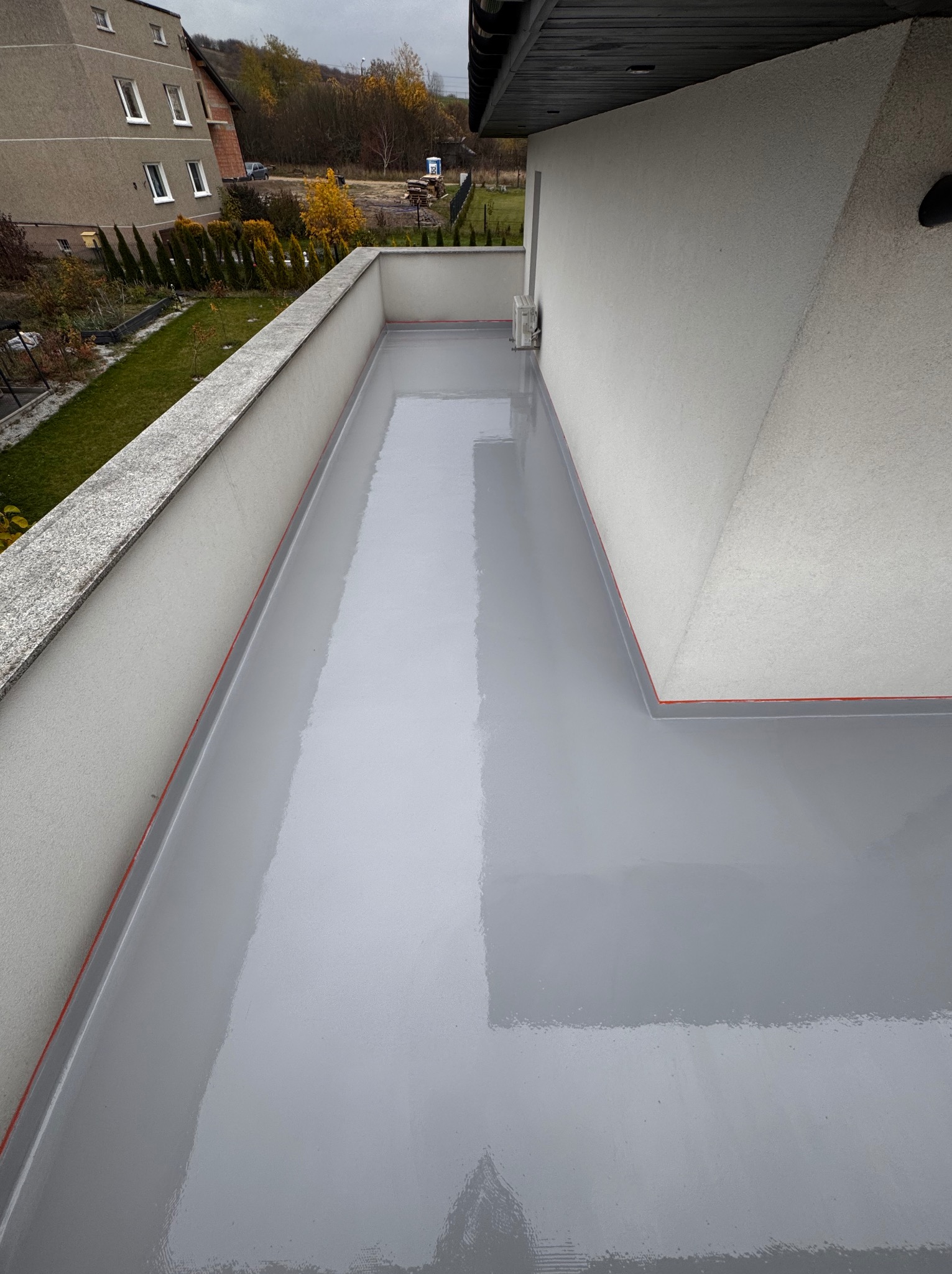 Szara, gładka powierzchnia balkonu z widoczną czerwoną taśmą zabezpieczającą przy ścianie, w tle budynek mieszkalny i drzewa.