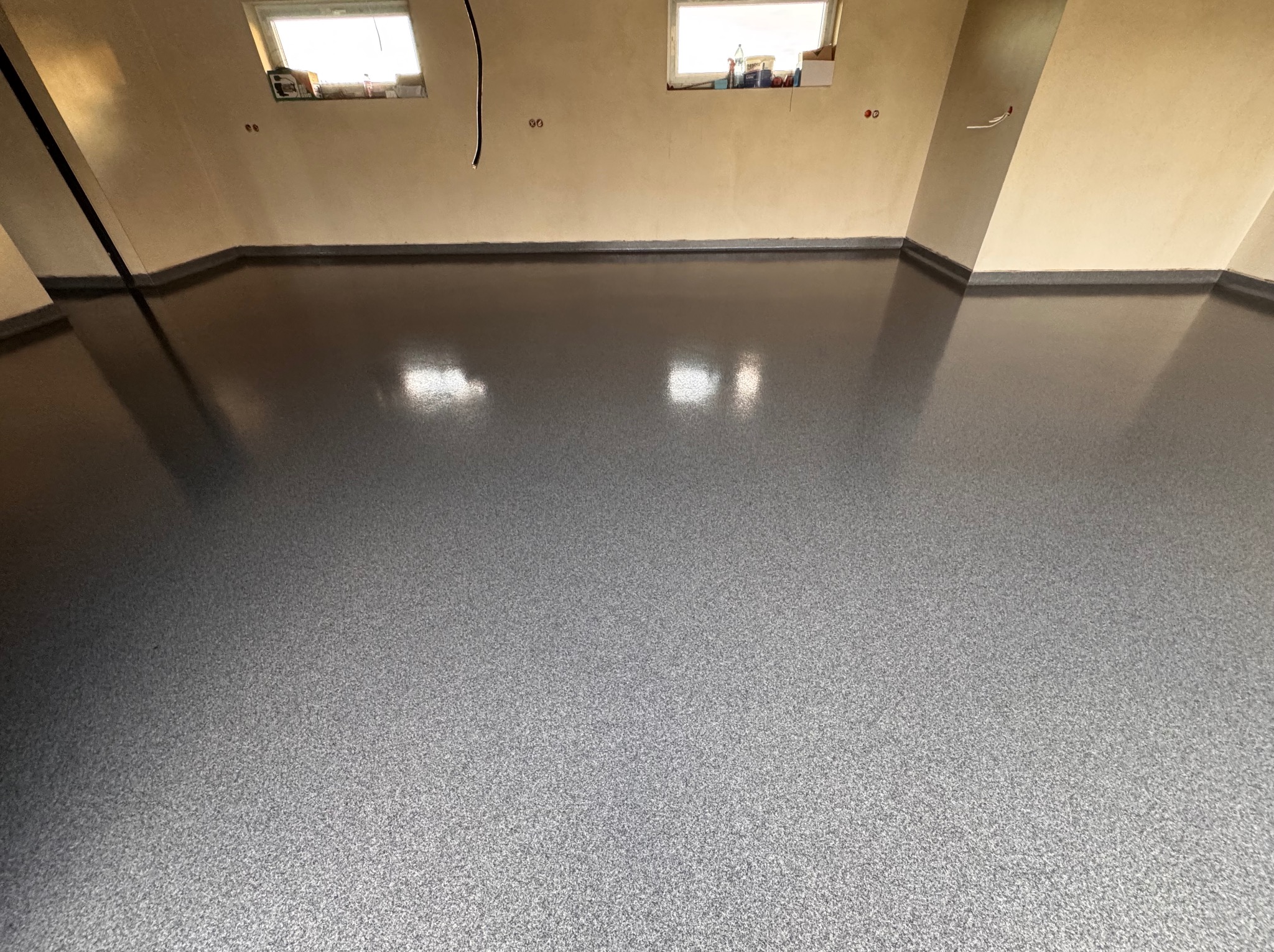 Szara, błyszcząca posadzka żywiczna w pomieszczeniu z beżowymi ścianami i dwoma oknami, widoczne kable elektryczne wystające ze ściany.