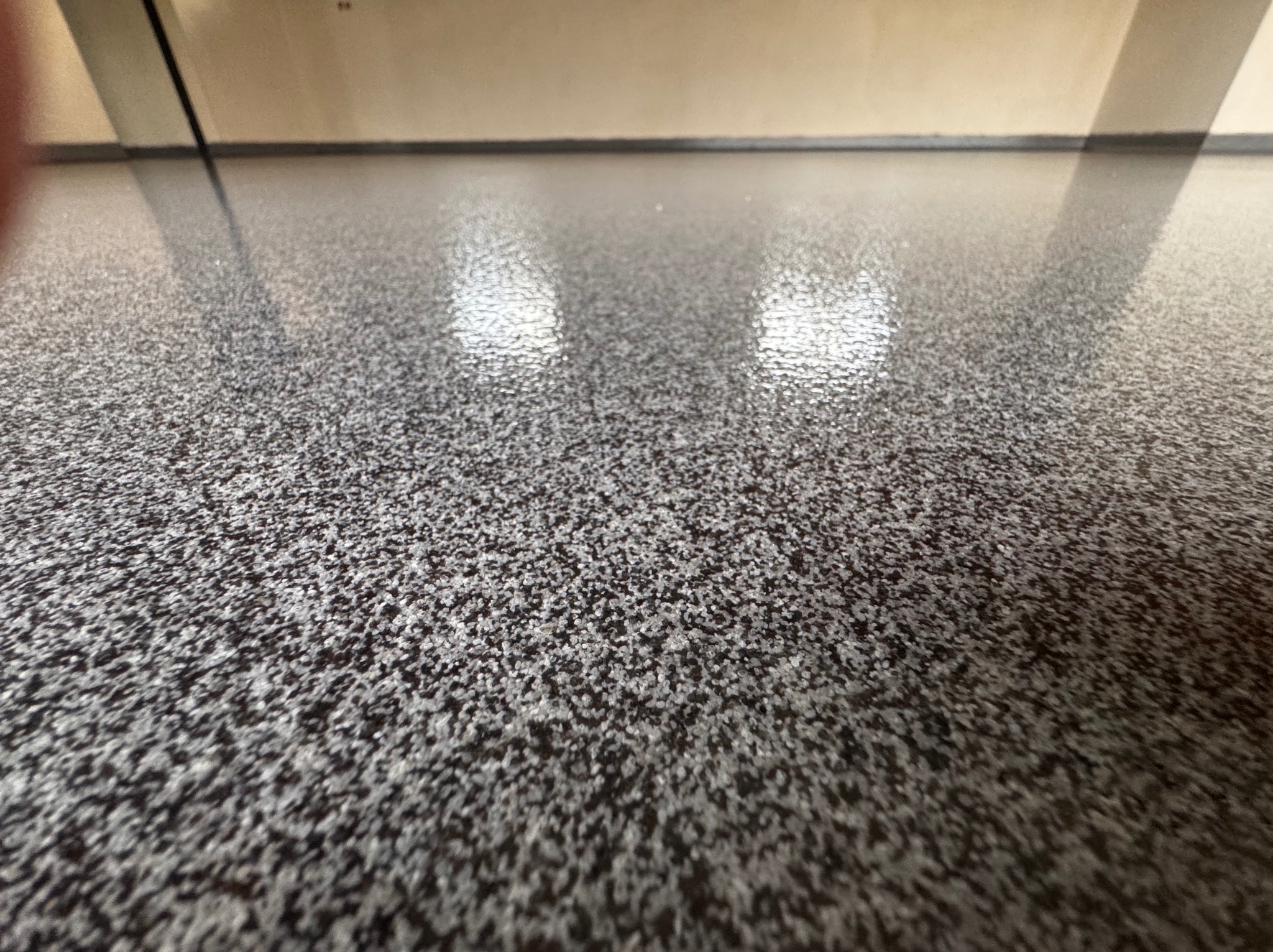 Szara, błyszcząca posadzka z drobnymi, ciemnymi i jasnymi elementami, widoczne odbicia światła.
