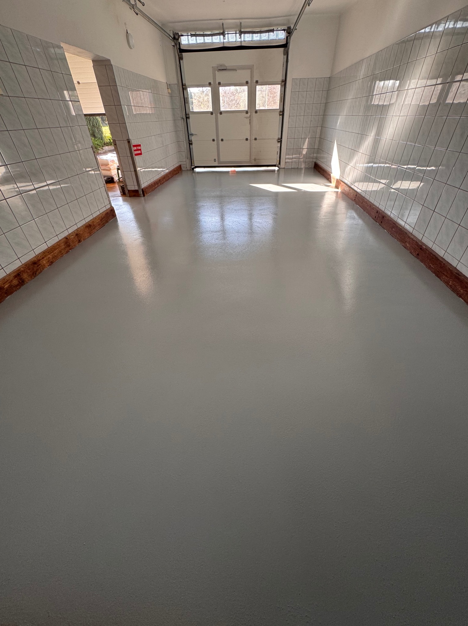 Jasnoszara, gładka posadzka w pomieszczeniu z białymi, kafelkowymi ścianami i drewnianymi listwami przypodłogowymi, światło słoneczne wpadające przez otwarte drzwi garażowe.