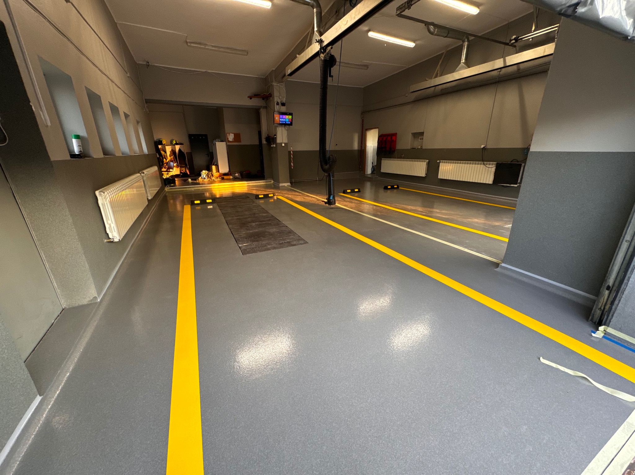 Szara posadzka w warsztacie samochodowym z żółtymi liniami wyznaczającymi stanowiska, widoczne kanały rewizyjne i system odciągu spalin.