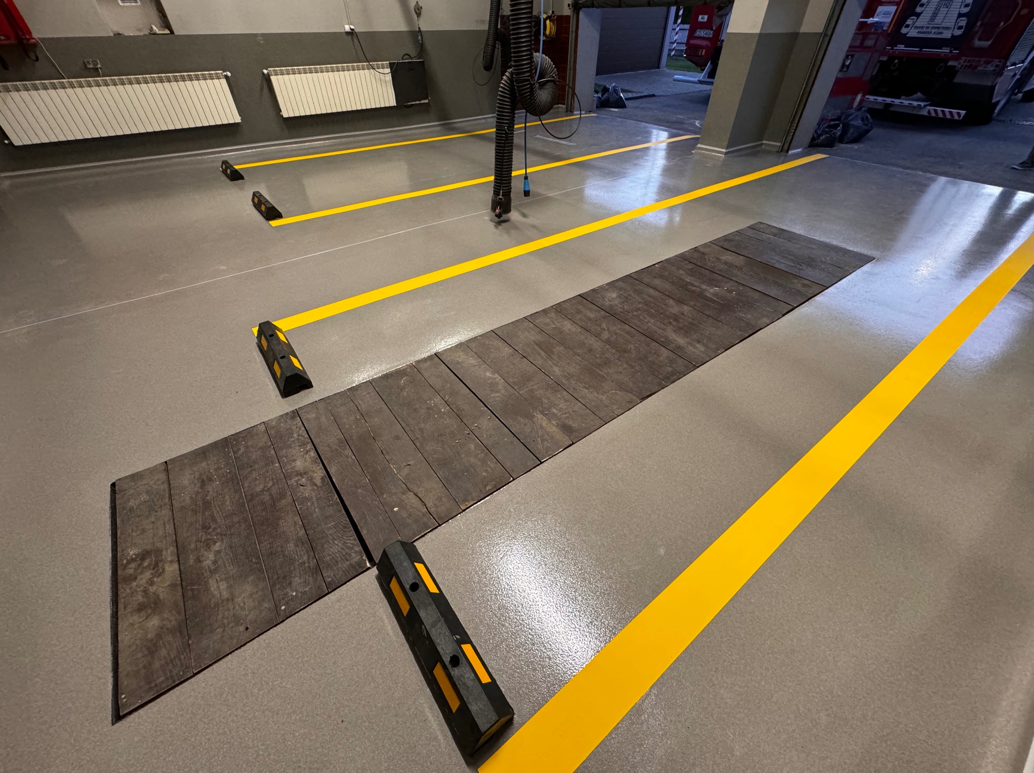 Szara posadzka w warsztacie z żółtymi liniami wyznaczającymi stanowiska, drewnianą platformą i odbojnikami przy stanowiskach.
