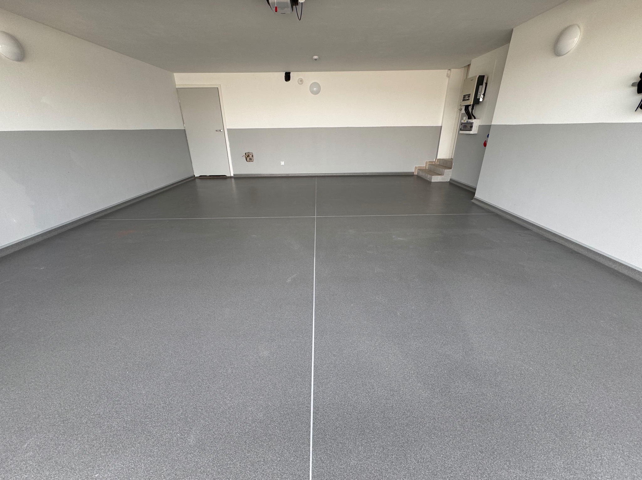 Szara, jednolita posadzka w garażu z widocznymi liniami podziału, ściany pomalowane dwukolorowo na biało i szaro, widoczne schody i instalacje elektryczne.