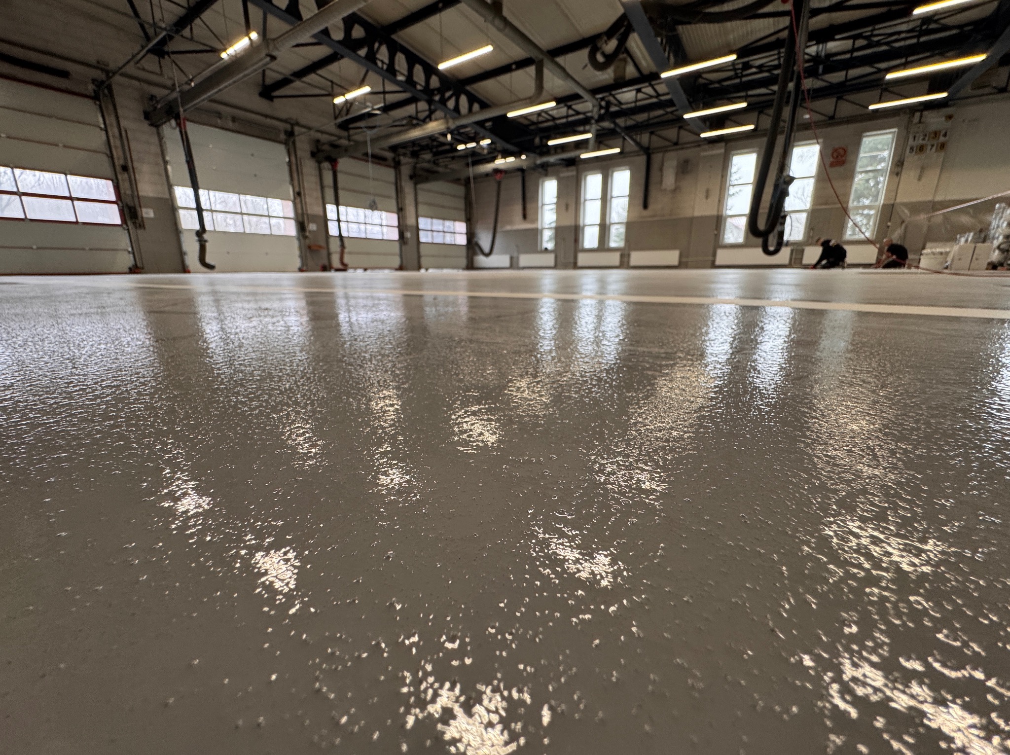 Gładka, szara posadzka w hali garażowej z widocznymi refleksami światła. W tle brama garażowa i okna. Dwóch pracowników w oddali.