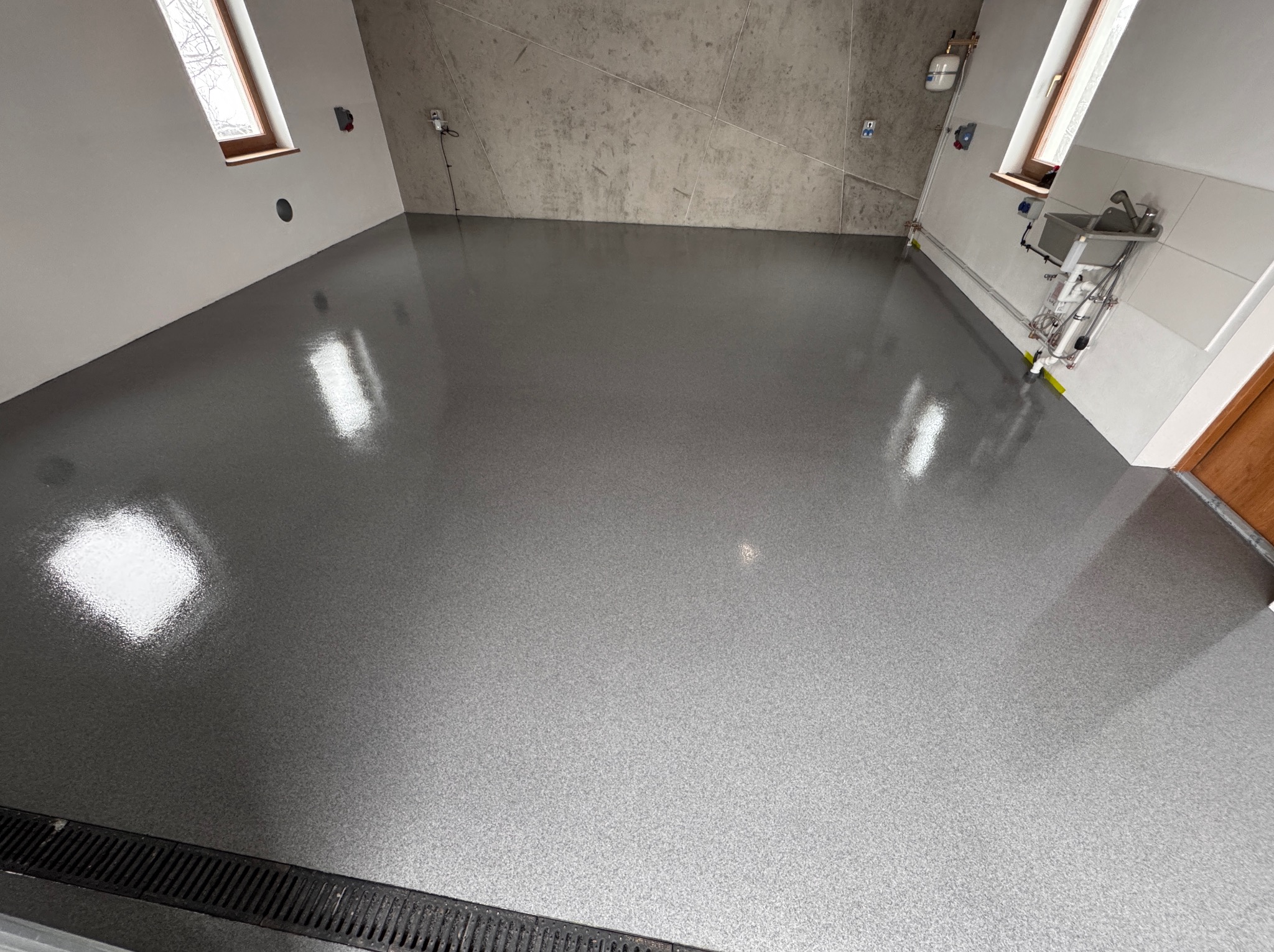 Błyszcząca, szara posadzka żywiczna z delikatnym wzorem w pomieszczeniu z oknami i instalacją wodną przy ścianie. Widoczny odpływ liniowy.
