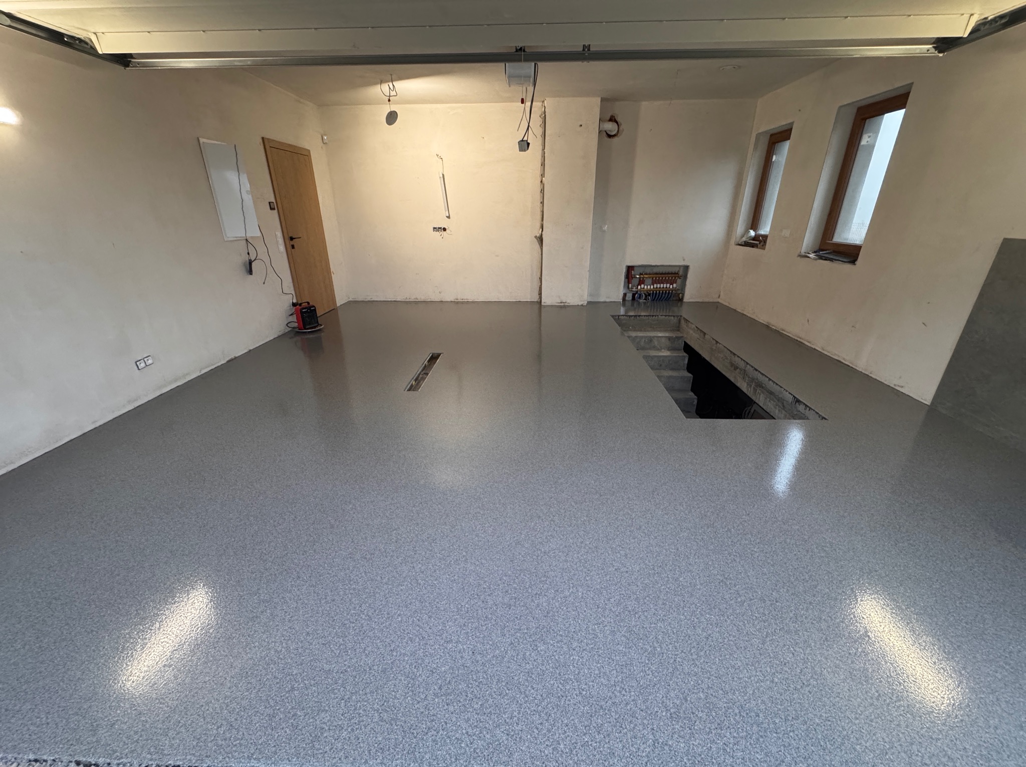 Szara, błyszcząca posadzka w garażu z kanałem i schodami. Widoczne okna i instalacje na ścianach. Nowoczesne wykończenie podłogi w surowym wnętrzu.