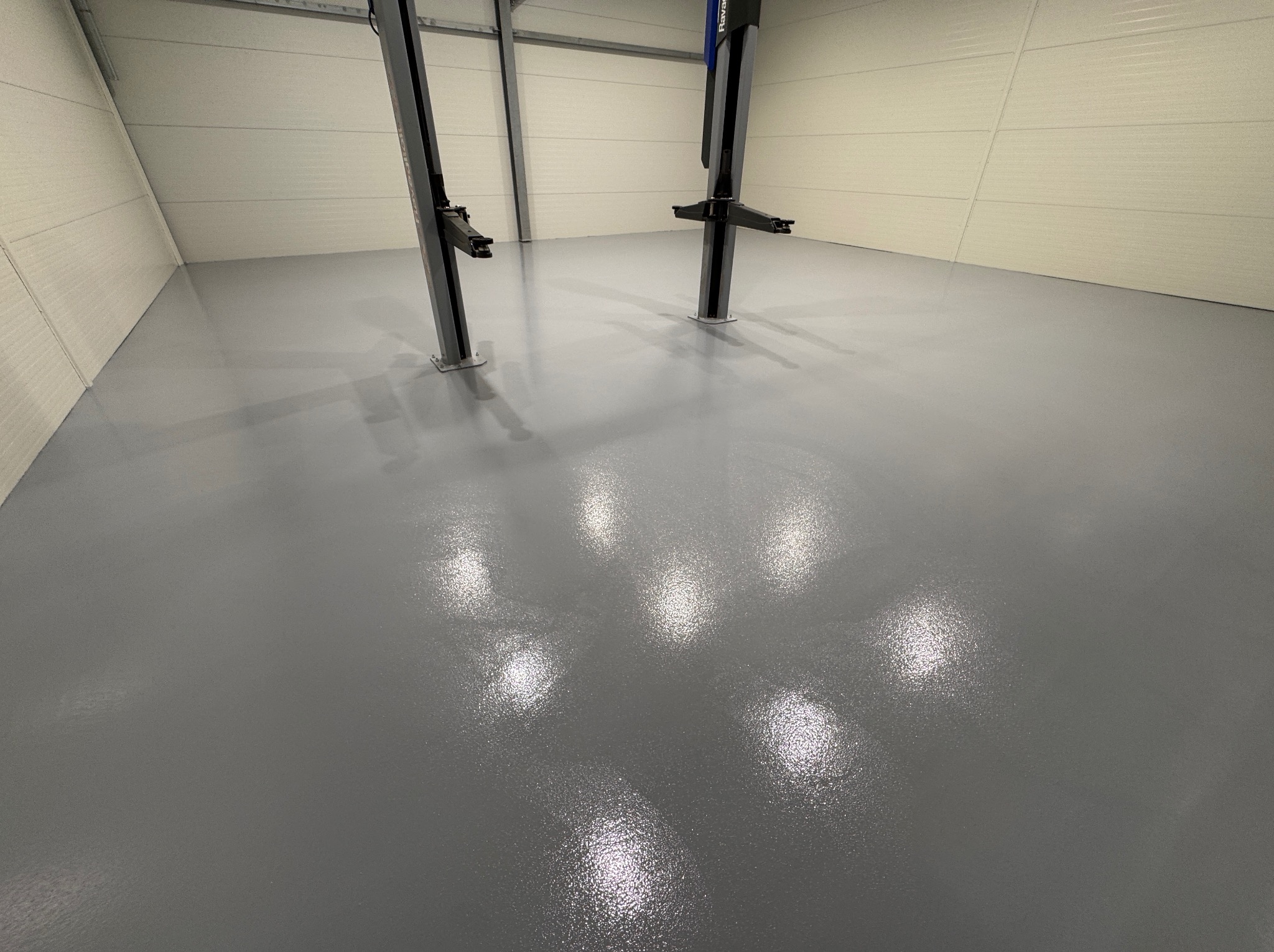 Gładka, szara powierzchnia w garażu z podnośnikami samochodowymi. Błyszcząca posadzka odbija światło, tworząc efekt lustrzany. Minimalistyczne wnętrze, widok z góry.