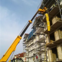 Żółta maszyna budowlana z wysięgnikiem teleskopowym i zsypem na gruz, pracująca przy elewacji budynku w trakcie budowy, rusztowania wzdłuż ściany, widoczni pracownicy.
