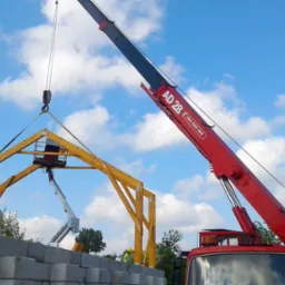 Czerwony dźwig AD 28 podnosi drewnianą konstrukcję dachu; w tle widoczny podnośnik koszowy i pracownik w żółtym kasku, a na pierwszym planie ułożone bloczki betonowe.