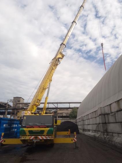 Żółty dźwig samochodowy z wysuniętym teleskopowym ramieniem na tle częściowo zachmurzonego nieba, z widocznym hakiem i fragmentem konstrukcji hali w tle.