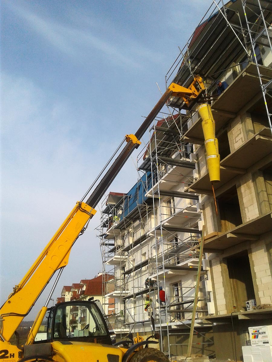Żółta maszyna budowlana z wysięgnikiem teleskopowym i zsypem na gruz, pracująca przy elewacji budynku w trakcie budowy, rusztowania wzdłuż ściany, widoczni pracownicy.