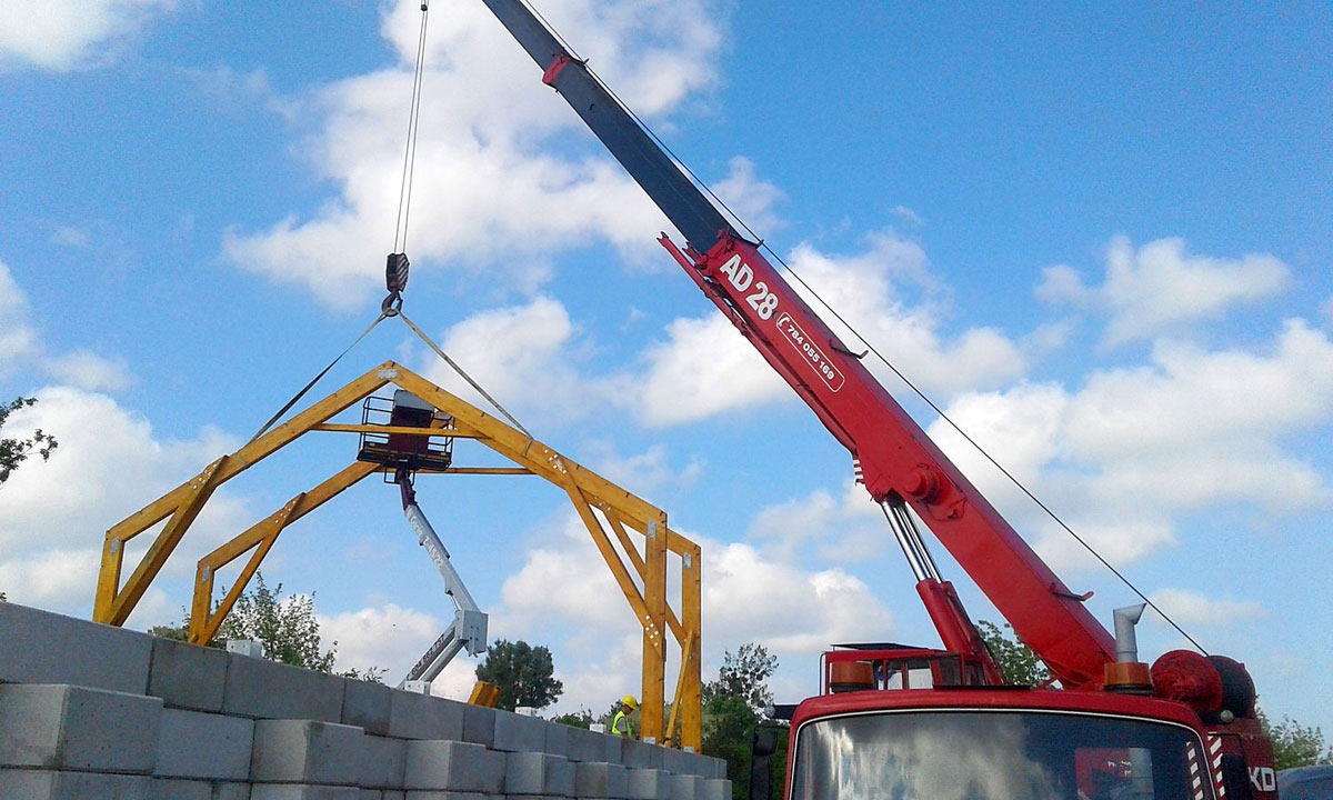 Czerwony dźwig AD 28 podnosi drewnianą konstrukcję dachu; w tle widoczny podnośnik koszowy i pracownik w żółtym kasku, a na pierwszym planie ułożone bloczki betonowe.