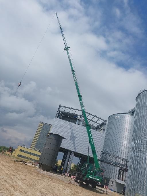Zielony dźwig teleskopowy w trakcie pracy na placu budowy, podnoszący element konstrukcyjny na tle srebrnych silosów i błękitnego nieba z chmurami.