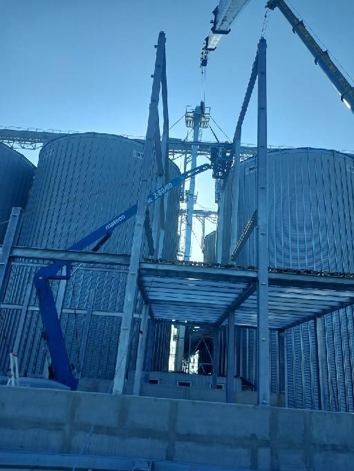 Montaż silosów zbożowych z użyciem żurawia i platformy roboczej na wysokości, błękitne niebo w tle.