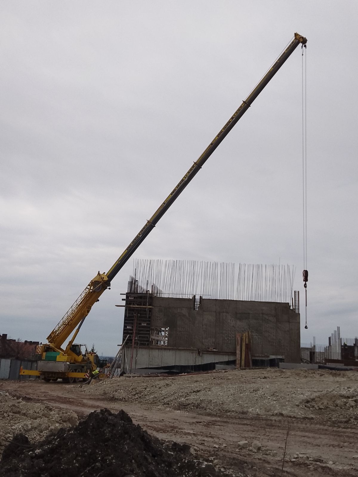 Żółty dźwig samochodowy z wysięgnikiem w trakcie pracy na placu budowy; widoczne zbrojenia i betonowe elementy konstrukcyjne budynku.
