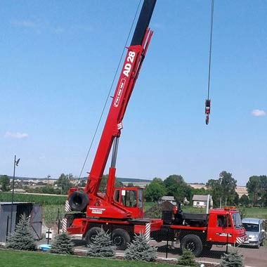 Czerwony dźwig samochodowy AD 28 na tle zielonego krajobrazu, z wysuniętym ramieniem i hakiem gotowym do podnoszenia, widoczny niewielki budynek gospodarczy i samochód osobowy w tle.