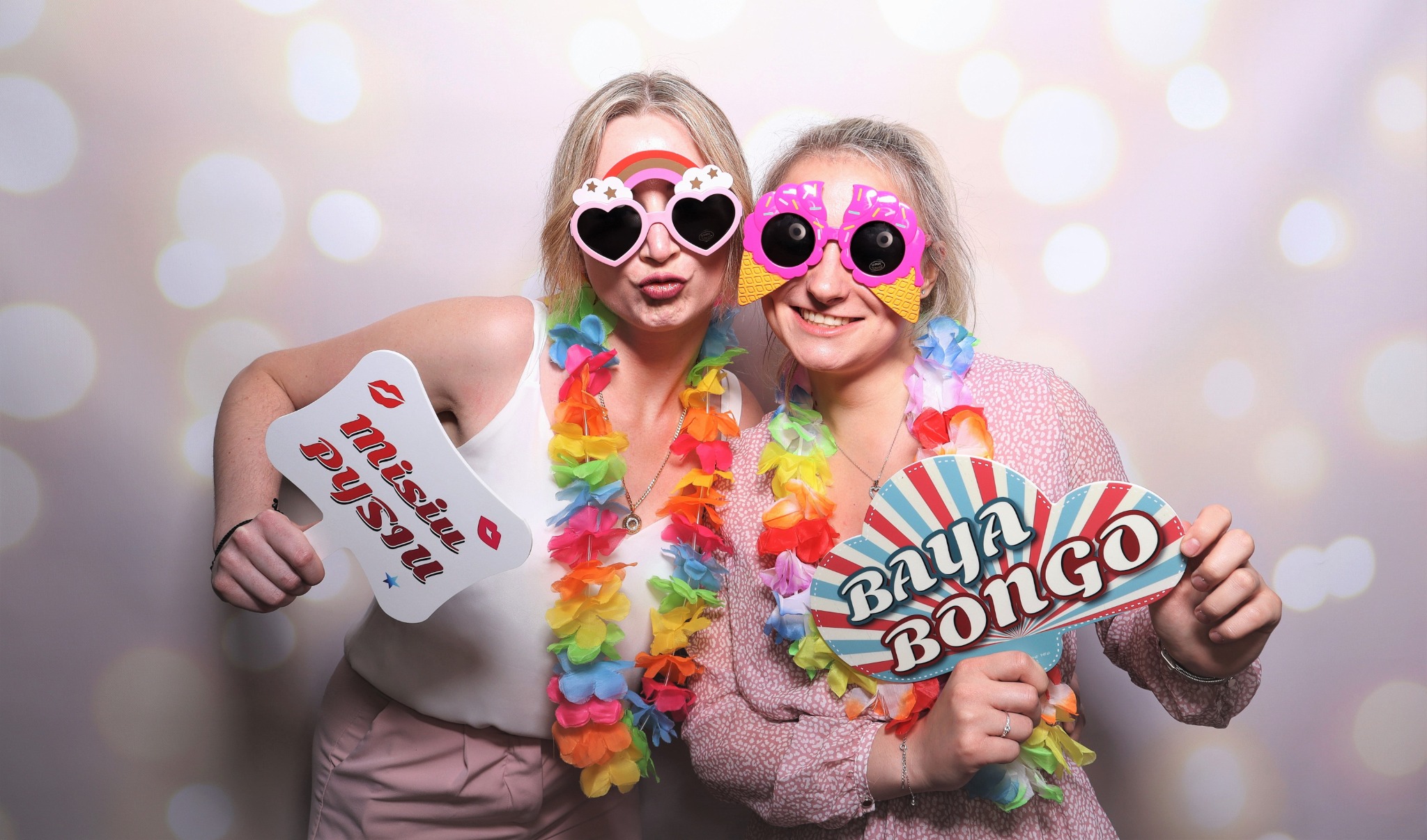Dwie uśmiechnięte kobiety w zabawnych okularach i hawajskich naszyjnikach pozują z rekwizytami 'Misiu Pysiu' i 'Baya Bongo' na tle światełek bokeh.