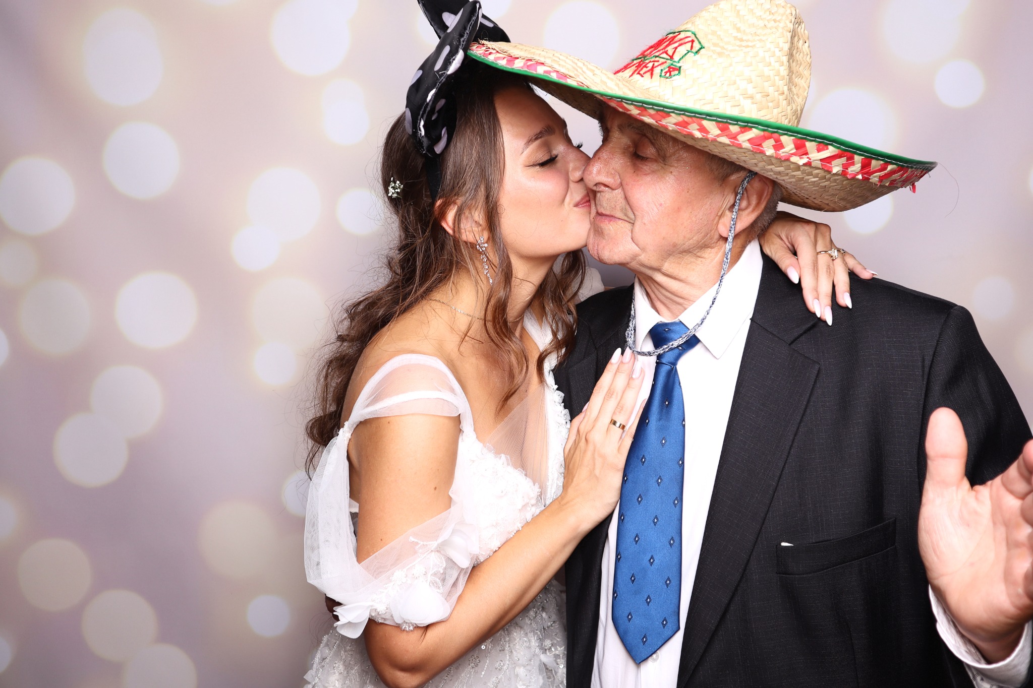 Uśmiechnięta kobieta w białej sukni całuje w policzek starszego mężczyznę w garniturze i meksykańskim sombrero na tle bokeh, zdjęcie wykonane w budce fotograficznej.