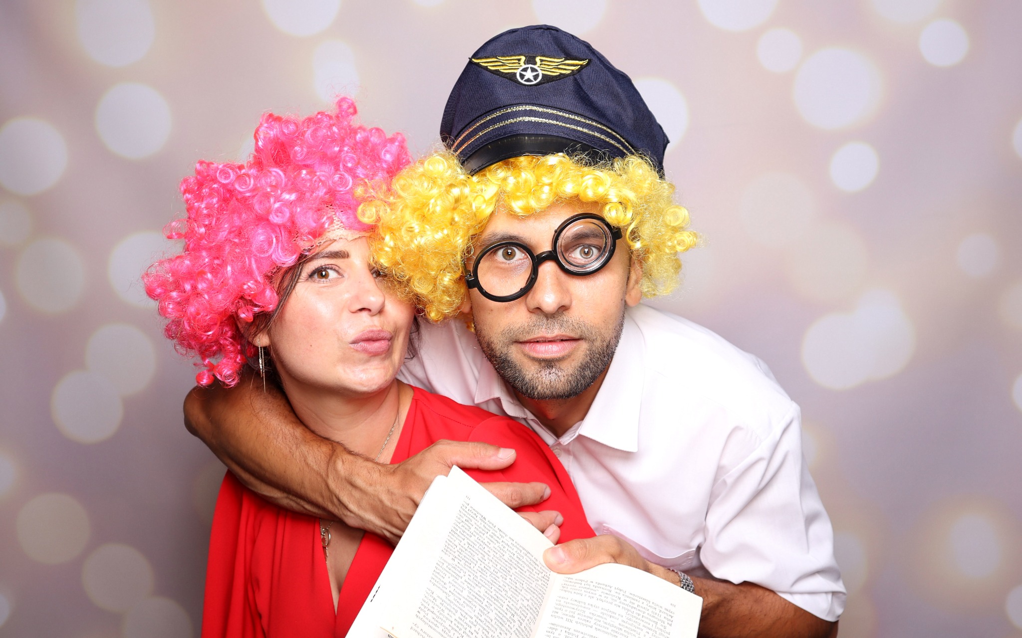 Para w przebraniach: kobieta w czerwonej sukience i różowej peruce, mężczyzna w peruce blond, czapce pilota i okularach, trzymający otwartą książkę, na tle studia fotograficznego z rozmytymi...