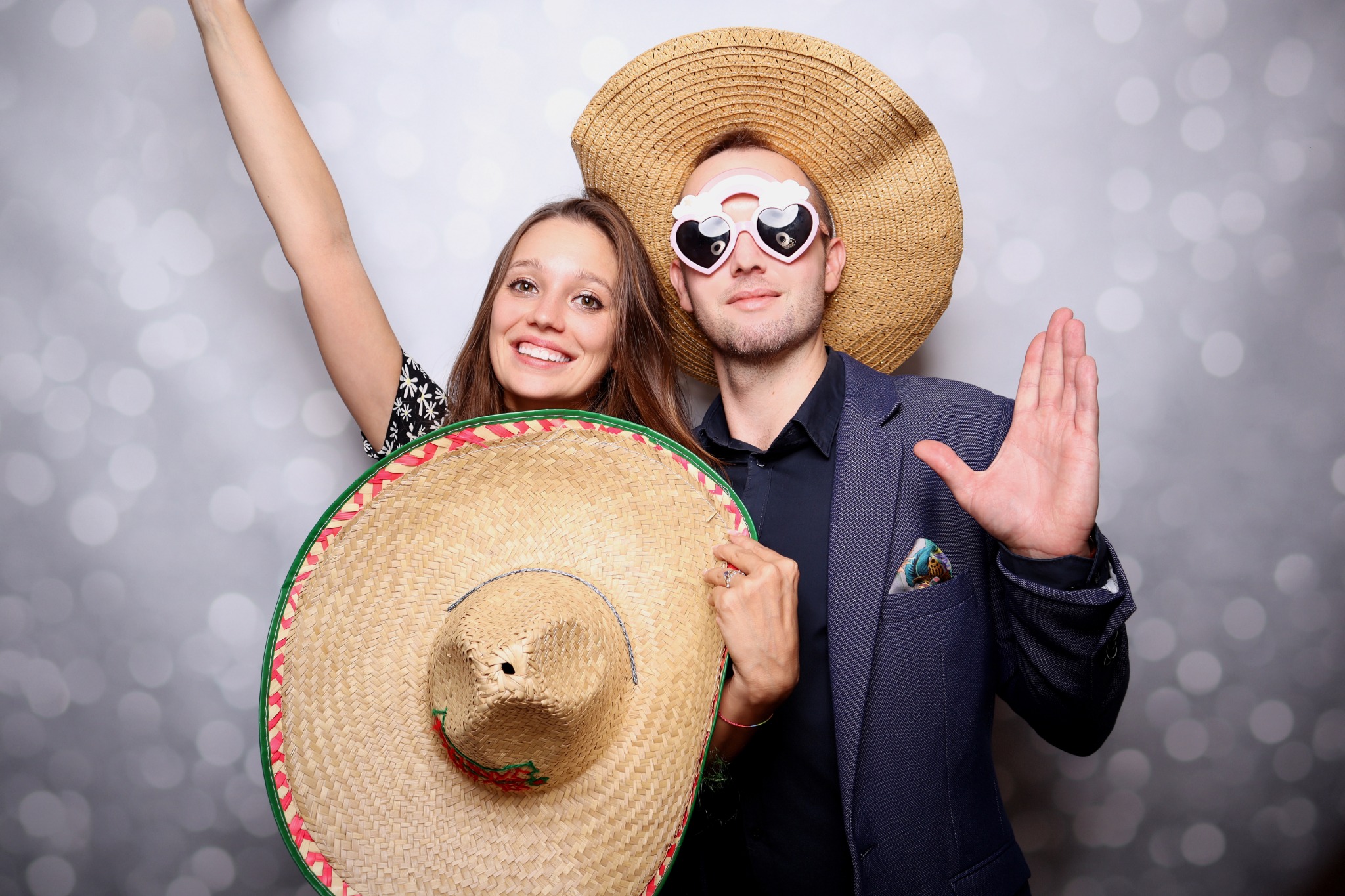 Para pozuje z rekwizytami: sombrero, duży słomkowy kapelusz i okulary w kształcie serc, na tle światełek bokeh.