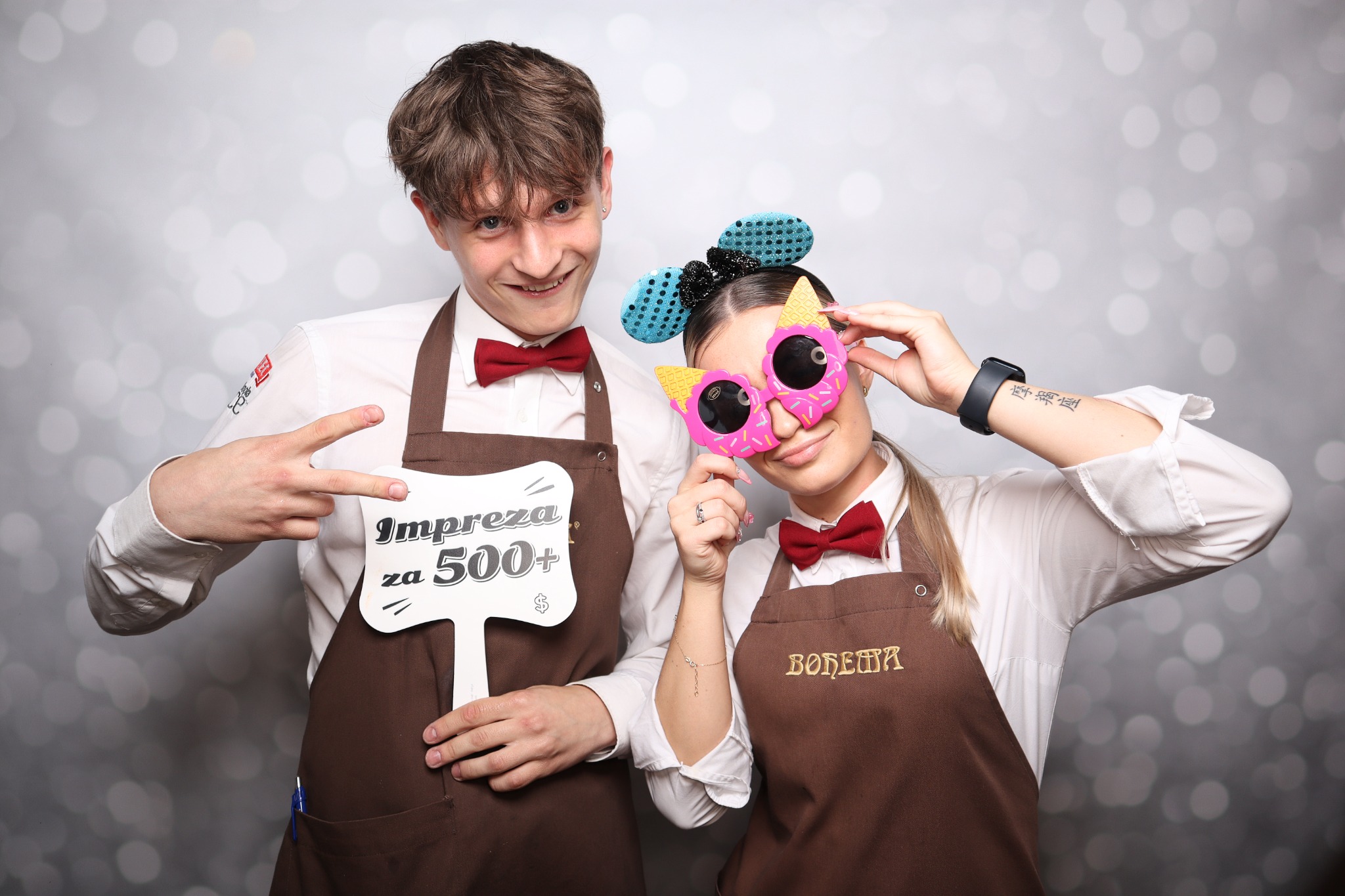 Dwie osoby, mężczyzna z napisem 'Impreza za 500+' i kobieta w okularach w kształcie lodów, pozują w brązowych fartuchach i czerwonych muchach na tle srebrnego, błyszczącego tła.