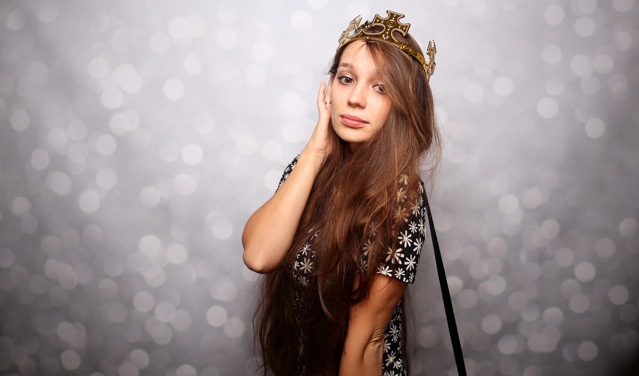 Portret kobiety w koronie na tle błyszczącego tła, z delikatnym uśmiechem i dłonią przy twarzy.