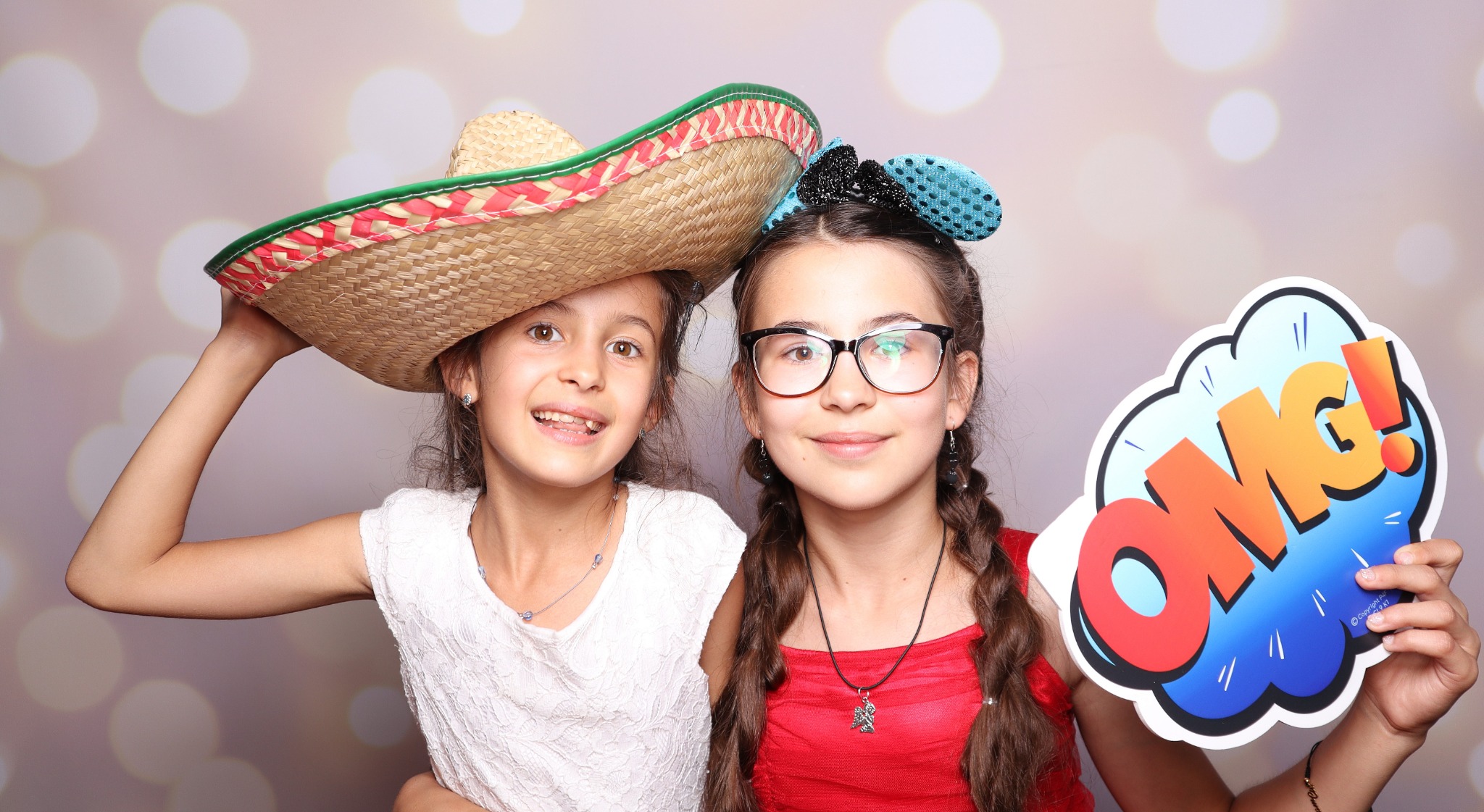 Dwie uśmiechnięte dziewczynki w fotobudce pozują z rekwizytami: sombrero i dymkiem z napisem 'OMG!'