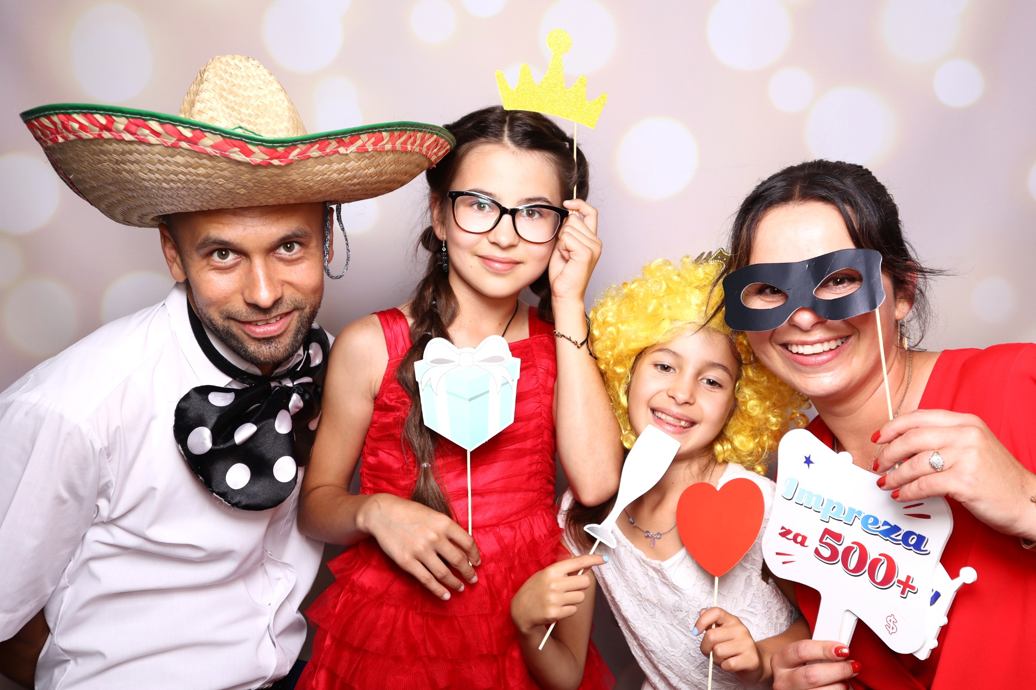 Uśmiechnięta rodzina pozuje z rekwizytami: sombrero, korona, maska karnawałowa, prezent, serce i tabliczka z napisem 'Impreza za 500+'.