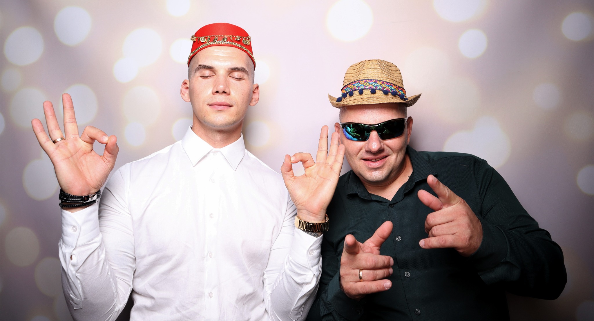 Dwóch mężczyzn pozuje na tle bokeh: jeden w fezie i białej koszuli z zamkniętymi oczami, drugi w kapeluszu, okularach przeciwsłonecznych i ciemnej koszuli, obaj pokazują gest 'OK'.