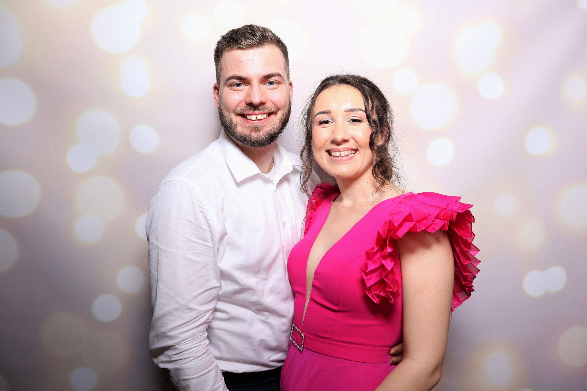 Para uśmiechnięta, pozująca na tle światełek bokeh, kobieta w różowej sukience z falbanami na ramionach, mężczyzna w białej koszuli.