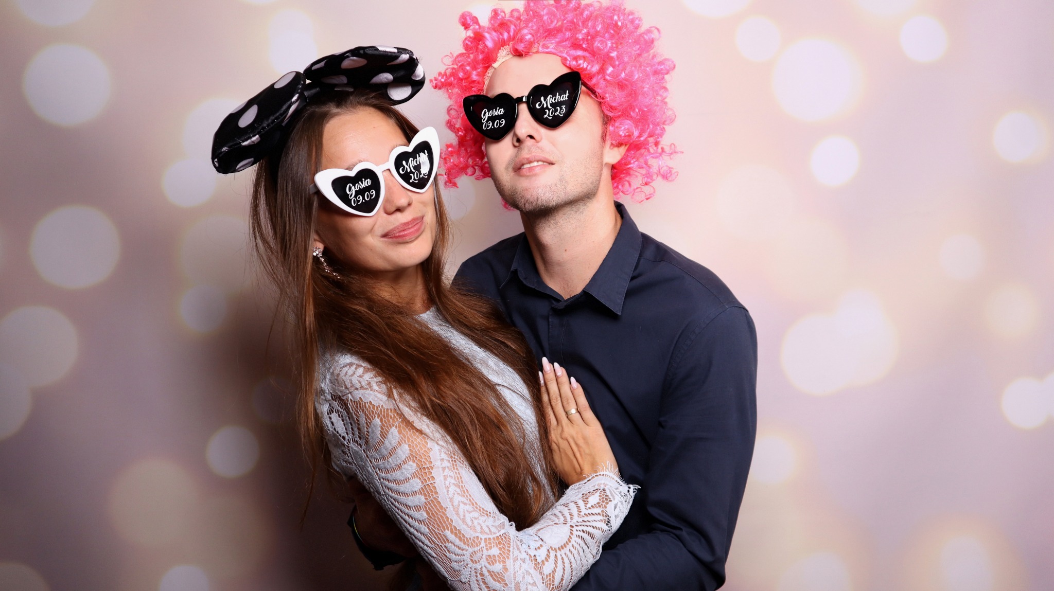 Para w okularach w kształcie serc z datami, kobieta z kokardą w kropki, mężczyzna w różowej peruce, pozują na tle bokeh.