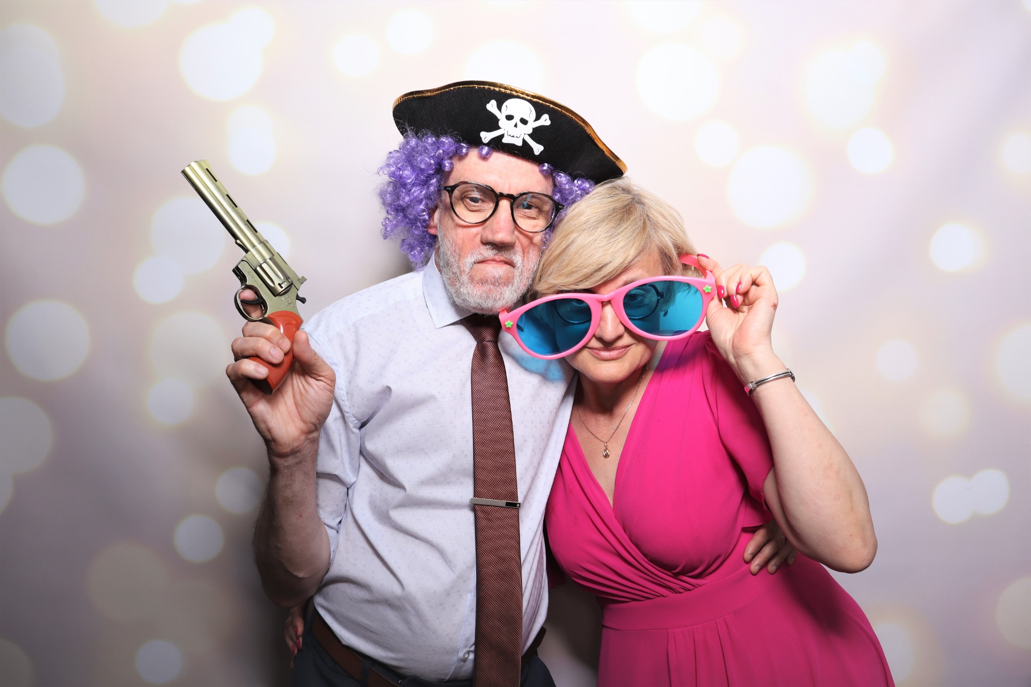 Para w przebraniach pozuje do zdjęcia w fotobudce we Wrocławiu: mężczyzna w pirackim kapeluszu i fioletowej peruce trzyma złotą zabawkę pistolet, a kobieta w różowej sukience ma na sobie duże...