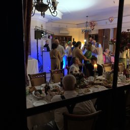 Twój Dj Maciej DJ SPIDER - Widok z okna na salę weselną: tańczący goście, DJ za konsoletą z niebieskim podświetleniem, eleganckie stoły z jedzeniem i świecami.