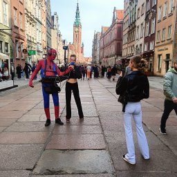 Twój Dj Maciej DJ SPIDER - Uliczny występ: Spiderman z mikrofonem i piosenkarką na tle zabytkowej ulicy Gdańska, nagrywani przez przechodnia. W tle tłum ludzi i wysoka wieża.
