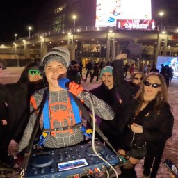 Twój Dj Maciej DJ SPIDER - DJ w stroju Spider-Mana, z mikrofonem i sprzętem DJ-skim, otoczony bawiącymi się ludźmi na tle budynku podczas nocnej imprezy plenerowej. Nietypowy strój i sceneria.