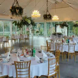 Elegancka sala bankietowa z okrągłymi stołami, złotymi krzesłami, kwiatowymi dekoracjami pod sufitem i oświetleniem girlandami. Widok na zewnątrz przez szklane ściany.