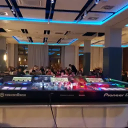 Pioneer DJ DDJ-RZX kontroler DJ-ski na stole z białym obrusem, w tle sala bankietowa z gośćmi i obsługą kelnerską, oświetlenie sufitowe w kolorze niebieskim.