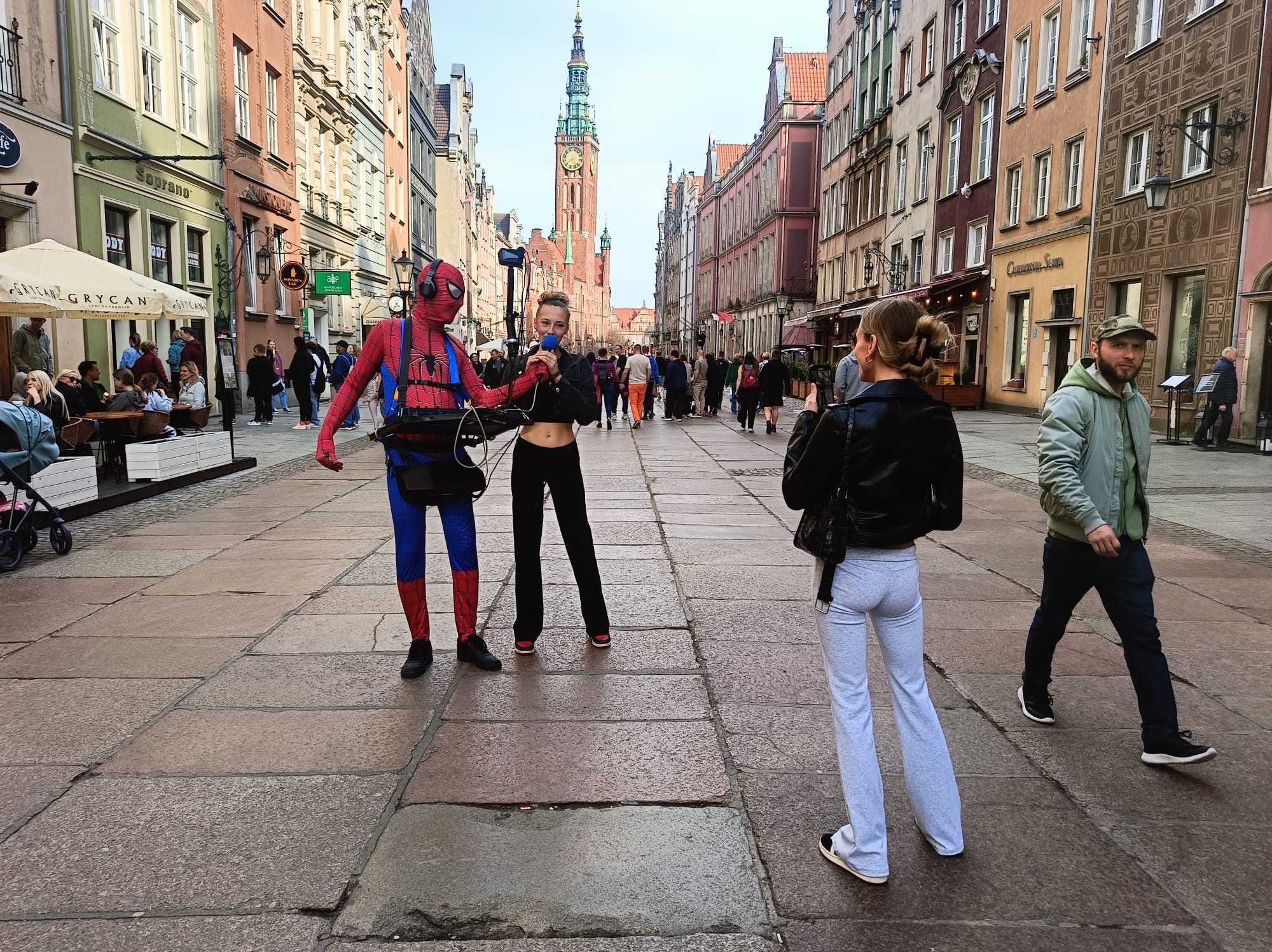 Uliczny występ: Spiderman z mikrofonem i piosenkarką na tle zabytkowej ulicy Gdańska, nagrywani przez przechodnia. W tle tłum ludzi i wysoka wieża.