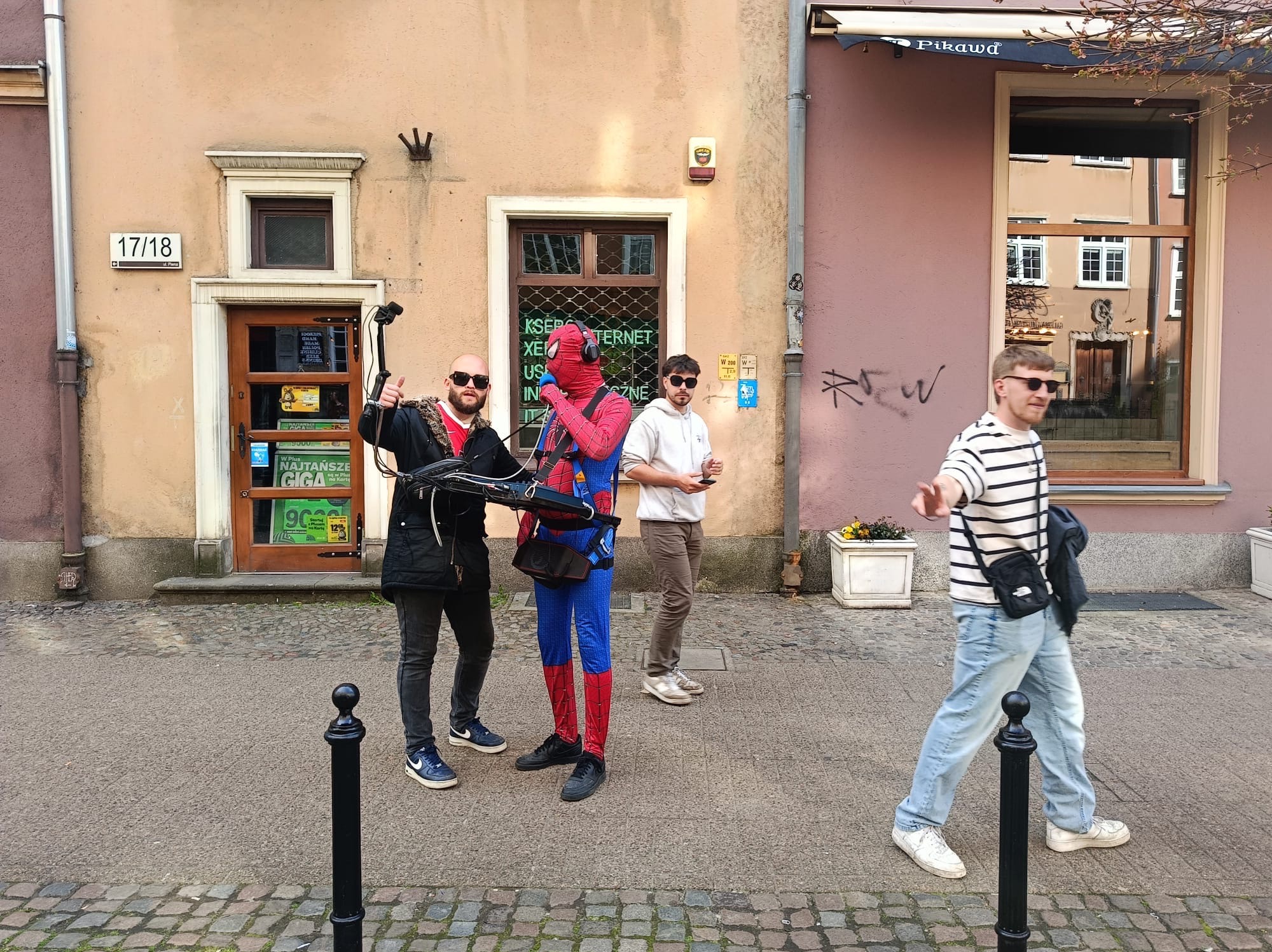 Ekipa eventowa w miejskiej scenerii: mężczyzna w kostiumie Spider-Mana, operator z kamerą i dwaj statyści na tle budynków. Nietypowe ujęcie z ulicy.