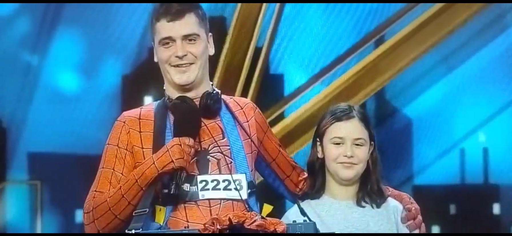 Animator w kostiumie Spider-Mana z mikrofonem i słuchawkami, pozujący z dziewczynką na tle sceny z niebieskim oświetleniem i złotymi elementami dekoracyjnymi.