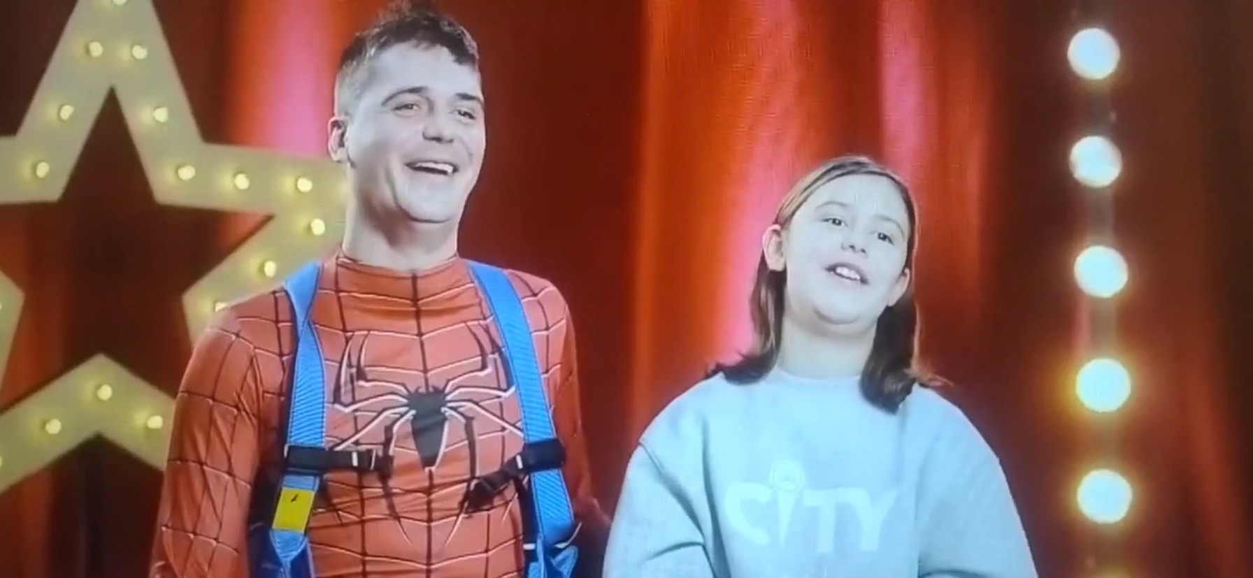 Animator w stroju Spider-Mana z uprzężą i dziewczynka na tle czerwonej kurtyny i gwiazd z żarówkami, uśmiechnięci, podczas występu dla dzieci.