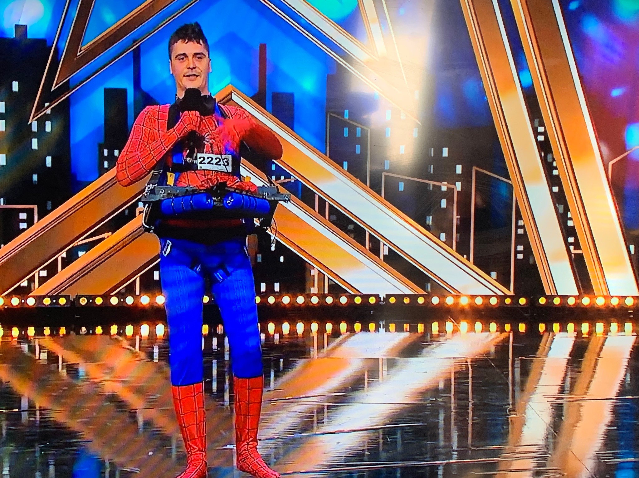 Mężczyzna w kostiumie Spider-Mana, z mikrofonem i rekwizytami, na scenie z oświetleniem i dekoracjami w geometryczne wzory. Numer startowy 2223.