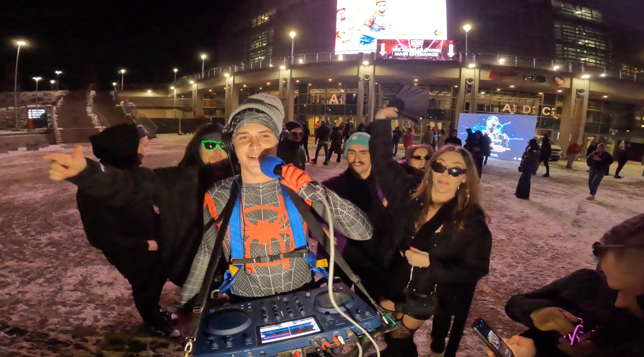 DJ w stroju Spider-Mana, z mikrofonem i sprzętem DJ-skim, otoczony bawiącymi się ludźmi na tle budynku podczas nocnej imprezy plenerowej. Nietypowy strój i sceneria.