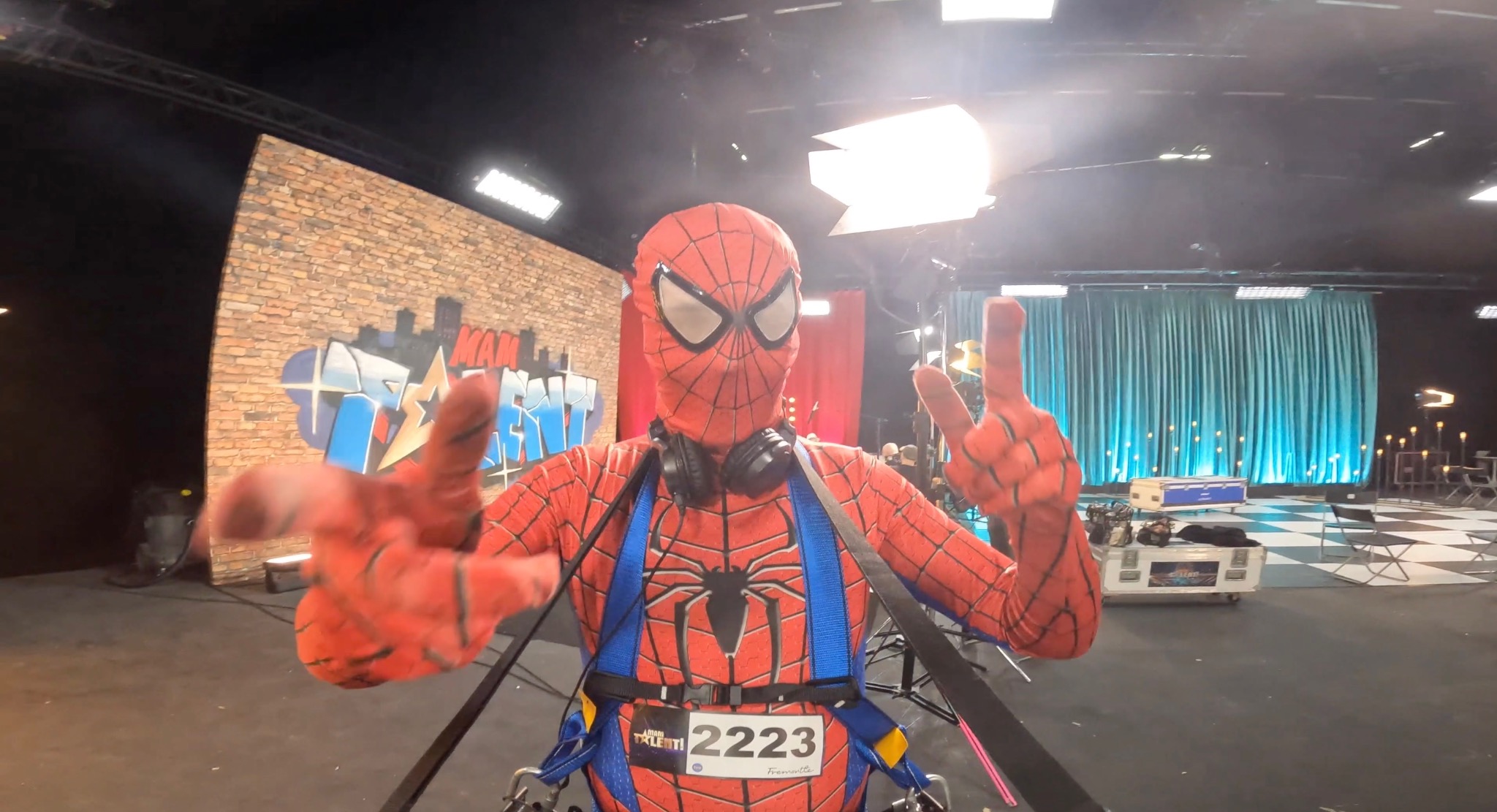 Animator przebrany za Spider-Mana na scenie z graffiti i turkusowymi zasłonami, pozujący z uniesionymi palcami w kształcie litery V. Na uszach ma słuchawki.