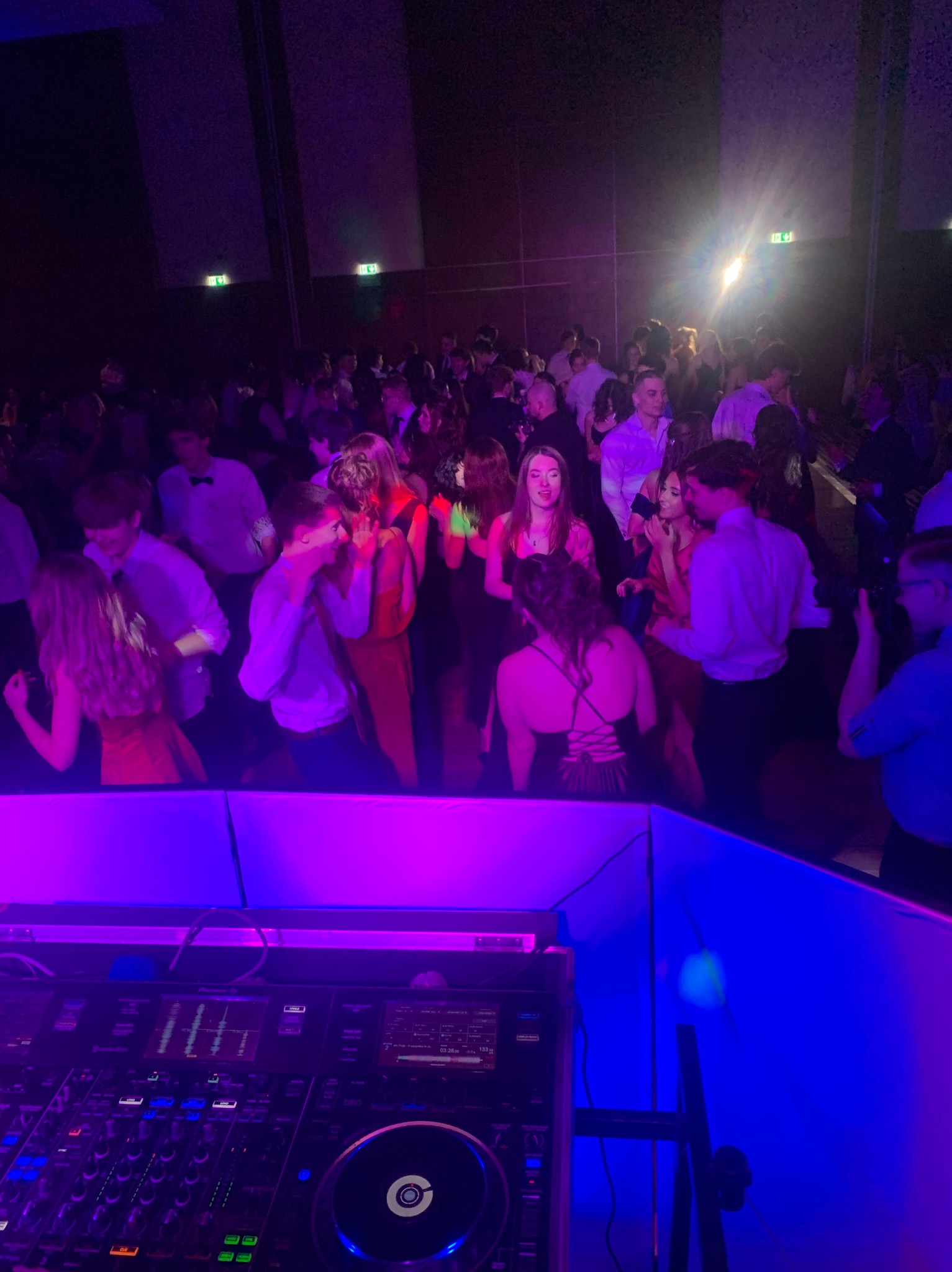 Parkiet taneczny wypełniony ludźmi podczas imprezy oświetlonej fioletowym i niebieskim światłem, widoczny sprzęt DJ-ski na pierwszym planie.