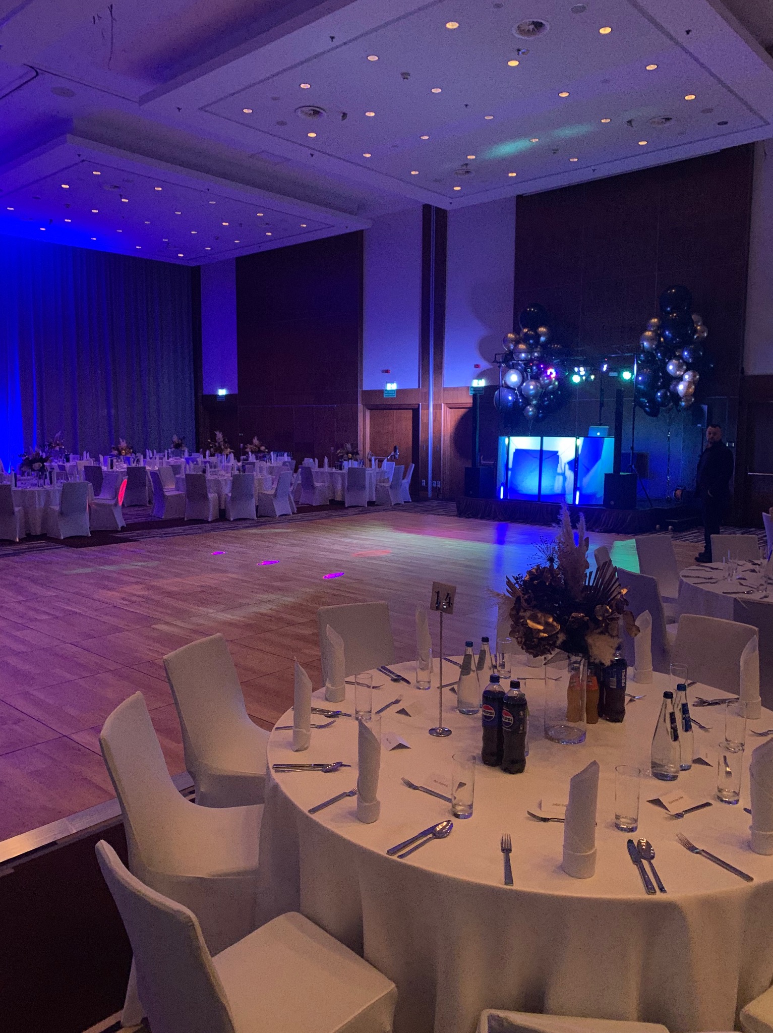 Sala balowa z okrągłymi stołami, dekoracjami kwiatowymi i sceną z nagłośnieniem. Przyciemnione światło, parkiet, przygotowana na imprezę. Dekoracje ze srebrnych balonów.